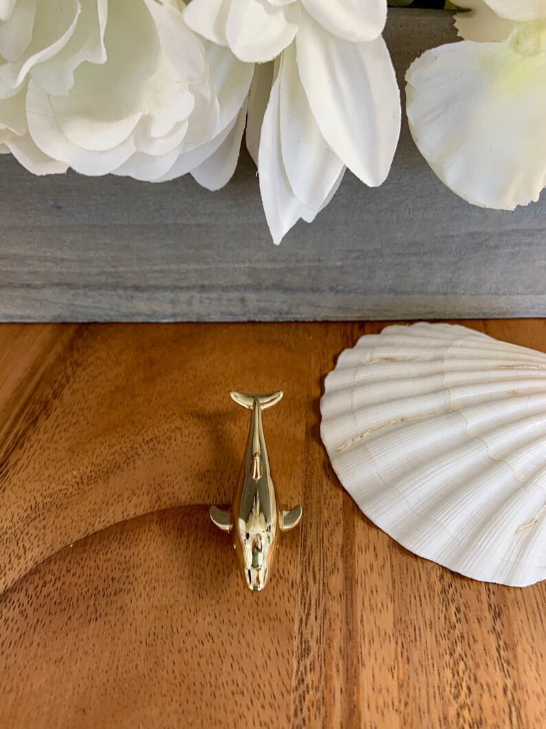 May include: A gold-colored whale figurine on a wooden surface. The whale is facing the camera with its tail up and its fins out. There is a white seashell to the right of the whale.
