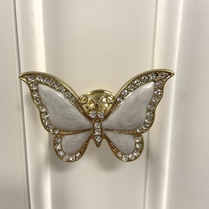 May include: A butterfly-shaped cabinet knob with a gold-tone metal frame, white marbled enamel wings, and clear crystal accents lining the edges of the wings and the center body. The hardware is mounted on a white paneled surface.
