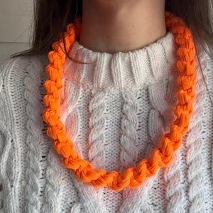 May include: Bright orange braided necklace. The necklace is made of a thick, woven material and is displayed against a white cable-knit sweater. The necklace is a bold statement piece, adding a pop of color to any outfit.