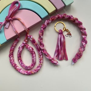 May include: Two pink and orange braided fabric headbands, one with a gold key ring and a rainbow charm. The headbands are on a white surface with a rainbow-colored wooden toy in the background.