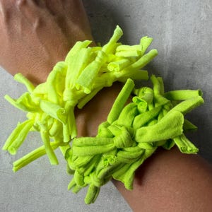 May include: Two knotted fabric bracelets made from strips of jersey material. One bracelet is a vibrant neon yellow, and the other is a bright lime green. Both feature a textured, shaggy appearance created by multiple tied fabric ends.