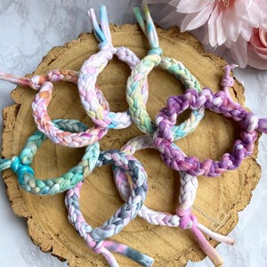 May include: A collection of seven braided fabric bracelets in a variety of pastel colors, including pink, purple, blue, and green. Each bracelet is tied at the ends. The bracelets are arranged on a wooden surface.