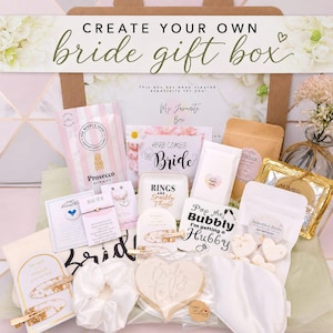 Pode incluir: Uma caixa de presente para noiva contendo vários itens, incluindo gomas de Prosecco, um prato para anéis com o texto Rings and Sparkly Things, um biscoito em forma de coração, um elástico de cabelo branco, ganchos, bombas de banho e cartões.