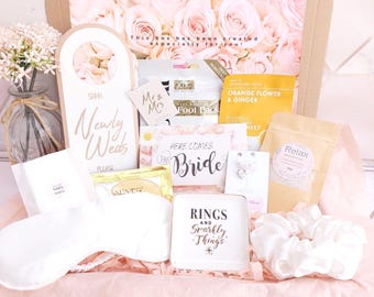 May include: A gift box containing a Newly Weds door hanger, a Here Comes The Bride card, a white sleep mask, a white scrunchie, a square dish labelled Rings and Sparkly Things, a Relax pouch, an Orange Flower & Ginger sheet, and a foot pack.