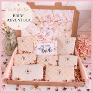 May include: A cardboard gift box containing seven numbered envelopes with beige patterns. The box lid features a floral rose print and text reading Create Your Own BRIDE ADVENT BOX. A central card displays the text HERE COMES THE Bride.