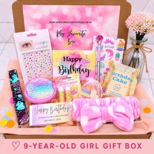 Pode incluir: Uma caixa de presente contendo adesivos faciais, um cartão de aniversário, um caderno, uma caneta com uma gema rosa, um saco de pipoca, uma faixa de lantejoulas, um bálsamo labial, uma barra de chocolate e uma faixa rosa de bolinhas.