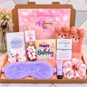 Pode incluir: Uma caixa de presente contendo uma máscara de dormir de pele sintética roxa, meias de urso felpudas cor-de-rosa, uma colher de chocolate quente com marshmallows, bombas de banho em forma de coração, um cartão de aniversário, uma pulseira e um elástico de cabelo rosa.