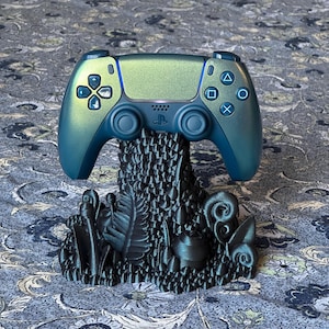 May include: A teal and blue video game controller rests on a dark teal decorative stand. The controller has a metallic finish and features the standard button layout. The stand is sculpted with a nature-inspired design, featuring leaves and other organic shapes.