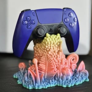 May include: A blue video game controller is sitting on a colorful 3D printed stand that resembles a fantastical plant or mushroom. The stand is a gradient of pink, orange, yellow, and green.