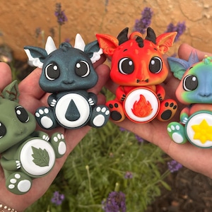 May include: Four small, stylized dragon figurines with large eyes and rounded features. Each dragon has a circular emblem on its chest: a green leaf, a black water droplet, a red flame, and a yellow star. The dragons are colored olive green, dark blue, bright red, and rainbow-patterned.