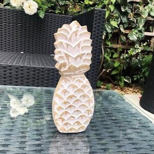 May include: White wooden pineapple sculpture with a detailed carved design. The sculpture is sitting on a black wicker surface.