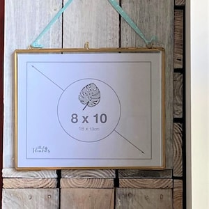 Puede incluir: Un marco de fotos rectangular dorado con un paspartú blanco, colgado de una cinta azul claro. El marco contiene una impresión con un diseño de hoja de monstera y el texto "8 x 10" y "18 x 13cm". El marco está montado sobre un fondo de madera rústica.