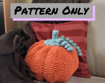 Può includere: Un cuscino a forma di zucca in crochet di colore arancione con uno stelo e foglie di colore verde turchese. Il cuscino è appoggiato su un divano con una coperta di lana marrone e un cuscino a righe rosse e bianche. Il testo "PATTERN ONLY" è visualizzato su uno stendardo grigio.