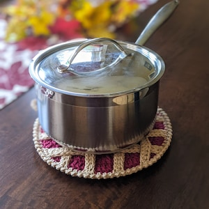 May include: A stainless steel pot with a lid and handle sits on a crocheted pot holder with a beige and burgundy design.