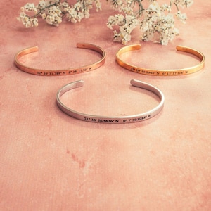 Puede incluir: Tres pulseras de oro y plata con coordenadas grabadas. Las pulseras est&aacute;n sobre un fondo rosa.