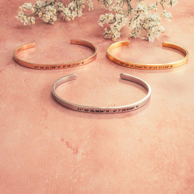 Puede incluir: Tres pulseras de oro y plata con coordenadas grabadas. Las pulseras est&aacute;n sobre un fondo rosa.