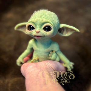 May include: A green figurine of a character with large black eyes and pointed ears. The figure is light green with darker green shading and is sitting in a hand. The text "CREATURES IN MY BAG" is visible.