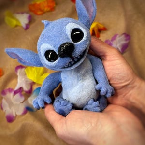 May include: A blue plush Stitch doll with large black eyes, a black nose, and a wide grin. The doll has large ears and small blue feet. The background includes colorful flower petals.