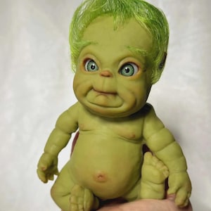 May include: A green baby doll with a green mohawk hairstyle and large green eyes. The doll is sitting with its legs crossed and its hands clasped together.