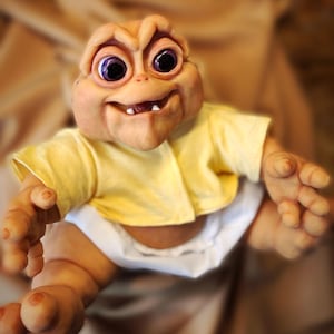 Pode incluir: Uma figura detalhada de um bebê dinossauro com uma expressão sorridente. A figura tem olhos grandes e roxos, uma camisa amarela e uma fralda branca. A pele do dinossauro é de cor castanho claro.