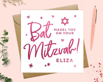 May include: A white Bat Mitzvah card with pink lettering and decorative hearts, stars, and a Star of David. The card reads "Bat Mitzvah!" and "Mazel Tov on Your" with the name "Eliza" below. A pink pen rests on the right.