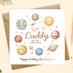 May include: A square greeting card with a white background featuring watercolour illustrations of smiling planets, a sun, a rocket, and a satellite. The text reads: TO THE BEST Daddy IN THE UNIVERSE and Happy birthday, love Amelia x in brown.