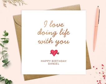 May include: A white greeting card with the message "I love doing life with you" in copper-colored script. A red heart pierced by an arrow is below the text, with "Happy Birthday Daniel" underneath. The card is on a brown envelope.
