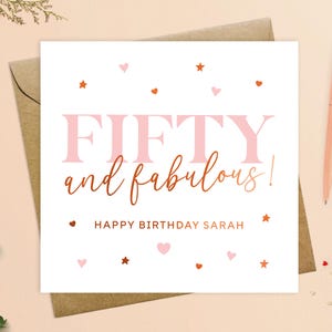 May include: A white card with pink and copper text that reads "Fifty and fabulous! Happy Birthday Sarah". The card is decorated with pink hearts and copper stars.