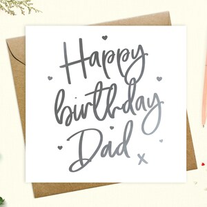 May include: A white card with silver lettering that says "Happy birthday Dad" with a small silver cross at the end of the text. There are small silver hearts scattered around the text.