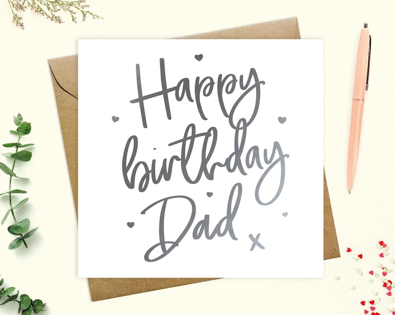 May include: A white card with silver lettering that says "Happy birthday Dad" with a small silver cross at the end of the text. There are small silver hearts scattered around the text.