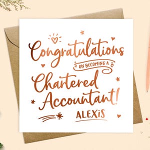 May include: A white card with copper-coloured text reading "Congratulations on Becoming a Chartered Accountant!" and the name "Alexis". The card is on a brown envelope, with a peach pen and greenery nearby.