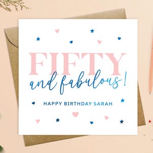 May include: A white card with pink and blue accents. The card says "Fifty and fabulous!" with the word "fifty" in pink and the words "and fabulous!" in blue. The card also says "Happy Birthday Sarah" in blue text. The card is decorated with pink hearts and blue stars.