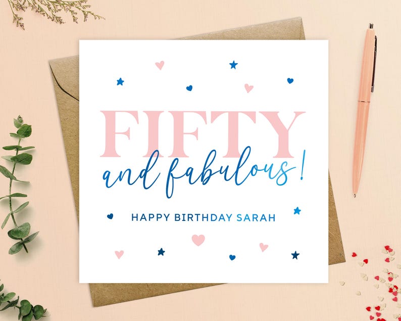 May include: A white card with pink and blue accents. The card says "Fifty and fabulous!" with the word "fifty" in pink and the words "and fabulous!" in blue. The card also says "Happy Birthday Sarah" in blue text. The card is decorated with pink hearts and blue stars.
