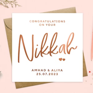 May include: A white card with a rose gold inscription that reads "Congratulations on your Nikkah" with two small hearts below the text. The names Ahmad & Aliya and the date 25.07.2023 are also written in rose gold below the hearts.