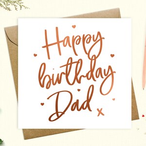 May include: A white card with a copper foil inscription that reads "Happy birthday Dad" with a small copper foil cross at the end of the inscription. There are small copper foil hearts scattered around the inscription.