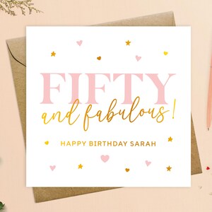 May include: A white card with pink and gold lettering that says "Fifty and fabulous!" with the name Sarah. The card is decorated with pink hearts and gold stars.
