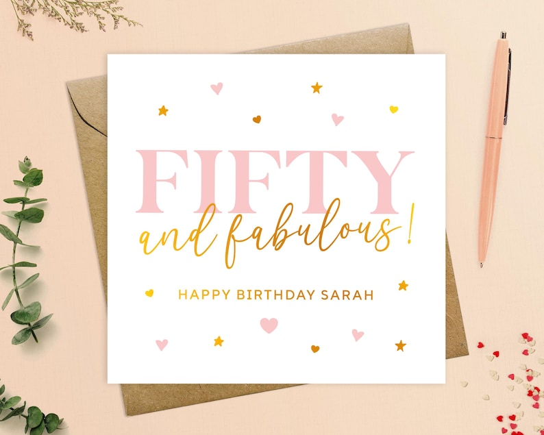 May include: A white card with pink and gold lettering that says "Fifty and fabulous!" with the name Sarah. The card is decorated with pink hearts and gold stars.