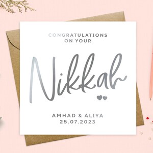 May include: A white card with silver foil lettering that says "Congratulations on your Nikkah" with two silver hearts below the word Nikkah. Below the hearts, it says "Ahmad & Aliya 25.07.2023".