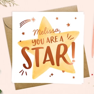 May include: A greeting card featuring a large yellow star and the words "Melissa, you are a STAR!" in brown lettering. The card is on a brown envelope, with a pink pen and decorative elements nearby.