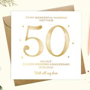 May include: A white card with gold lettering. The card says "To my wonderful husband Matthew" and "50" in large gold letters. The card also says "On our Golden Wedding Anniversary 14.02.2026" and "With all my love".