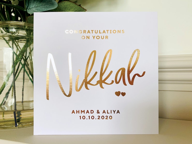 May include: White card with gold foil lettering. The card says "Congratulations on your Nikkah" with two small hearts below the word Nikkah. Below the hearts, it says "Ahmad & Aliya 10.10.2020".