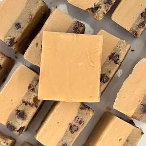 May include: A close-up of several squares of fudge topped with cookie dough. The fudge is a light brown color and the cookie dough is a darker brown color with chocolate chips.