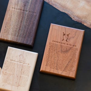 May include: Wooden plaques with engraved text from biblical verses. The plaques are rectangular and vary in wood tone, from light to dark brown. One plaque features the text "Philippians 2:1-11", another "Ephesians 6:10-20", and a third "Revelation 21:1-8".