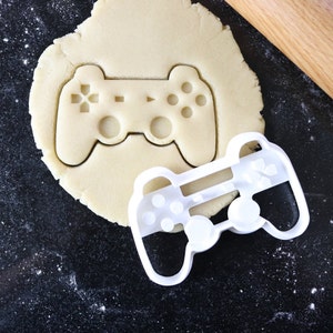 Peut inclure: Un emporte-pièce en plastique blanc en forme de manette de jeu vidéo. L'emporte-pièce est posé sur une pâte à biscuits ronde.