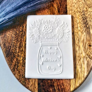 May include: White ceramic stamp featuring a design of a mason jar with sunflowers and the text "Happy Mother's Day".