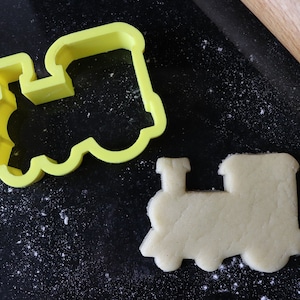 Peut inclure: Un emporte-pièce en plastique jaune en forme de locomotive, à côté d'un biscuit nature non cuit en forme de locomotive.