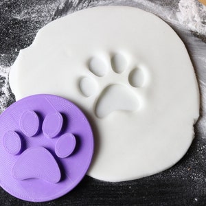 May include: A purple plastic paw print cookie cutter on a white surface with a paw print impression in the surface.