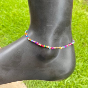 May include: A colorful beaded anklet with a variety of small, round beads in shades of blue, orange, red, green, and white. The anklet is displayed on a black mannequin foot against a backdrop of green grass.
