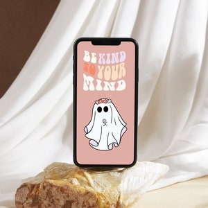 以下が含まれることがあります： 黒いスマートフォンは、「BE KIND TO YOUR MIND」というテキストが入ったピンク色の壁紙を表示しています。壁紙には、花の冠をかぶり、小さな花を持っている漫画の幽霊が描かれています。携帯電話は明るい色の岩の上に置かれています。