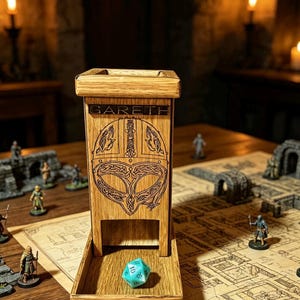 May include: A light brown wooden dice tower featuring a laser-engraved Viking helmet design and the name GARETH etched at the top. The tower has an open front tray where a single turquoise twenty-sided die rests on the flat wooden surface.